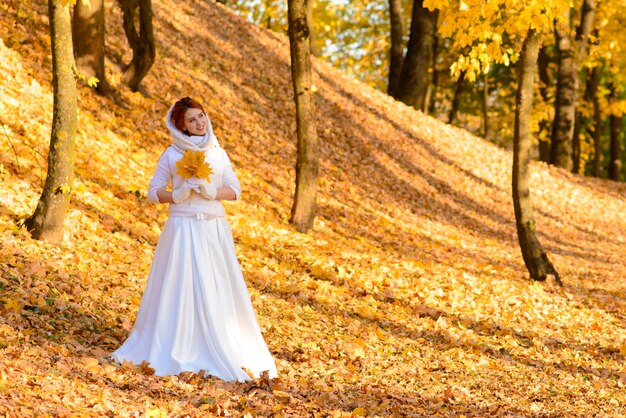 Cute girl in long white wedding dress posing in rural path among autumnal trees in forest