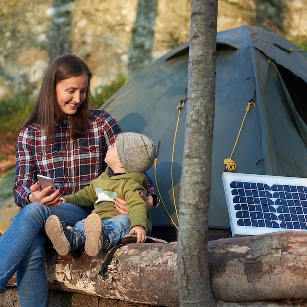 Cute girl looks at child sitting on log in forest near solar pan