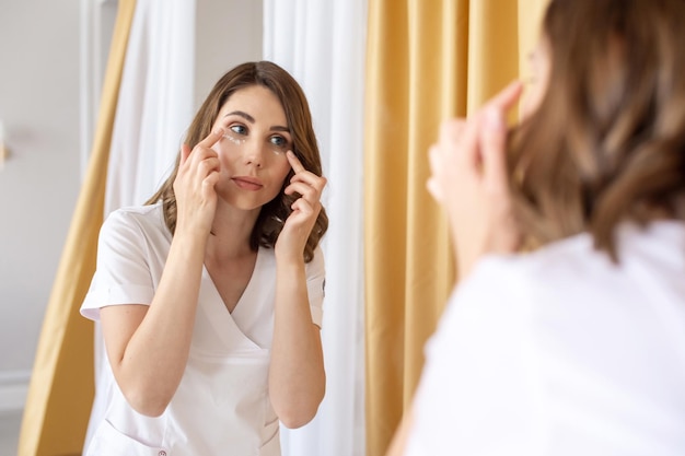 Foto una ragazza carina si guarda allo specchio e si applica la crema per gli occhi con punti bianchi