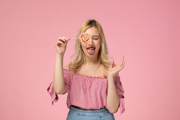 Cute girl lovely young lady with red lipstick in pink blouse covering one eye with lollipop