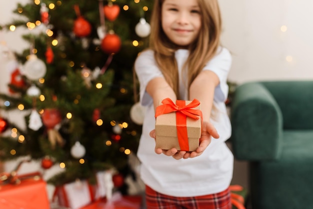 Cute girl in pajamas gives a gift to the camera on the background of the Christmas tree