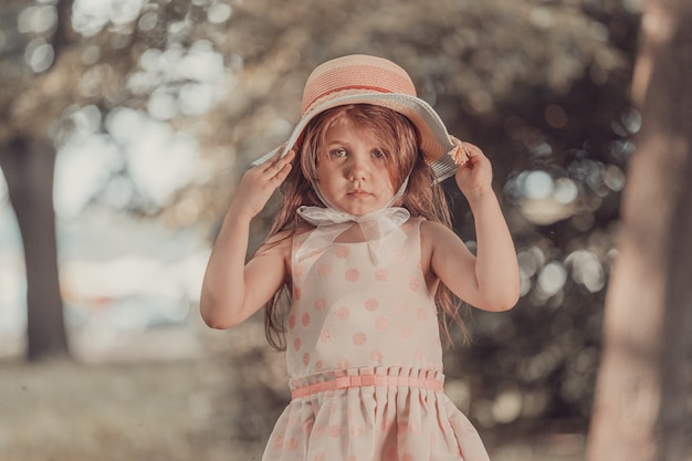 Милая девушка в розовом платье с шляпой стоит в парке летом. Фото высокого качества