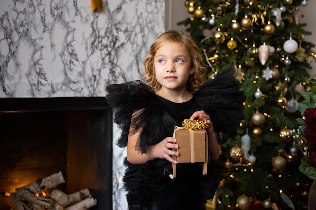 Photo cute girl playing with christmas present in her hands and christmas trees with lights merry christmas and happy holidays