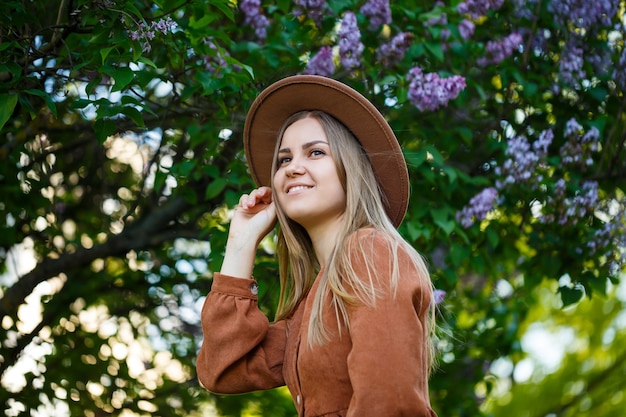 Cute girl portrait in brown big hat and stylish clothes against the background of a lilac tree in a botanical garden. Long light brown hair lies on the shoulders