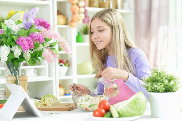 Cute girl preparing fresh salad on kitchen table with tablet at home