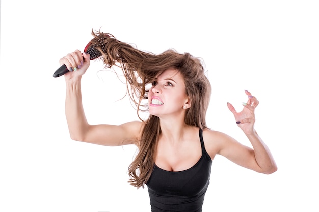 Cute girl pulls out a comb from tangled hair and screams into the camera on a white wall. Hair health concept.