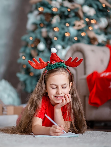 A cute girl in a Santa hat writes a letter to Santa near the Christmas tree Happy childhood