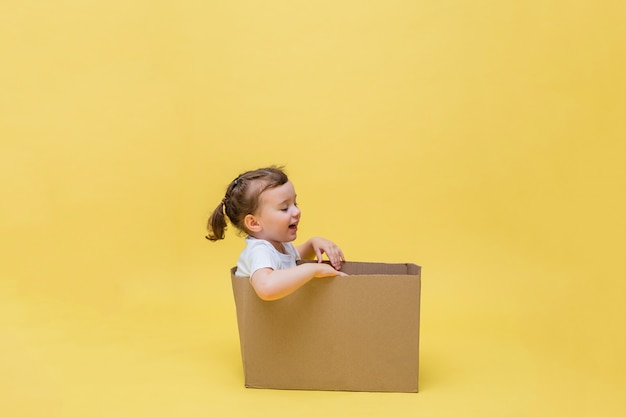 귀여운 소녀는 골판지 상자에 앉아 멀리 보인다. 노란색 공간에 흰색 티셔츠에 어린 소녀. 비접촉식 배송