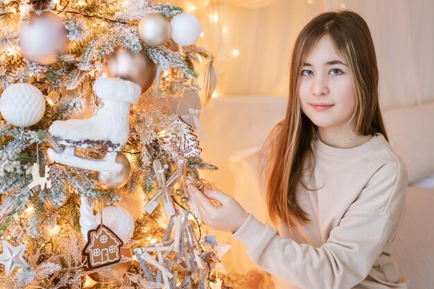 Cute girl sits on floor by window with christmas tree in background in her bedroom