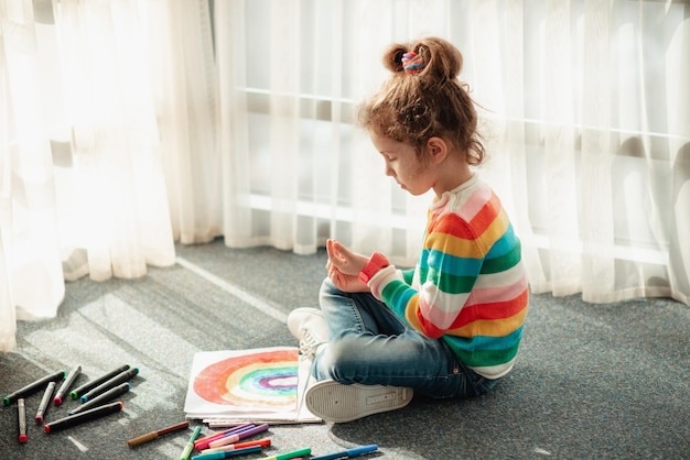 A cute girl sits on the floor in the rays of the sun and draws a rainbow with colorful markers