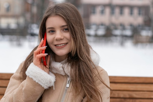 Cute girl sits on park bench and talks on phone with red case Smiling young woman in beige sheepskin coat rests outside in winter