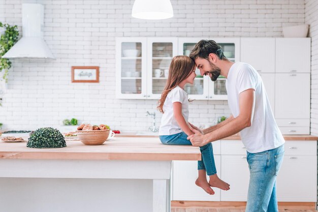 Cute girl sitting on the table and father standing on the kitchen and looking at each other side view