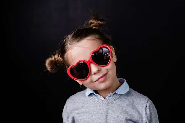 Cute girl in stylish sunglasses with heartshaped lens standing in front of camera in isolation against black space