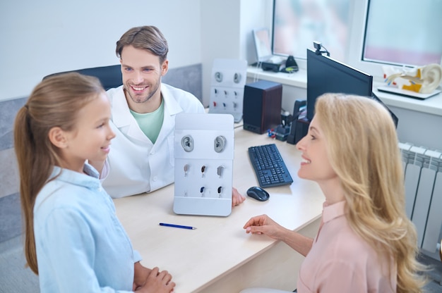 Cute girl trying on a new deaf aid in front of her smiling mother and a pleased male doctor