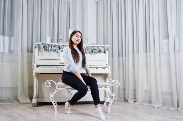 Cute girl wear on warm sweaters and black pants against old piano with christmas decoration at white room.