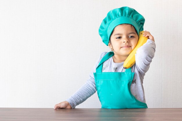 写真 シェフのユニフォームを着た可愛い女の子が家で壁にバナナで遊んでいます