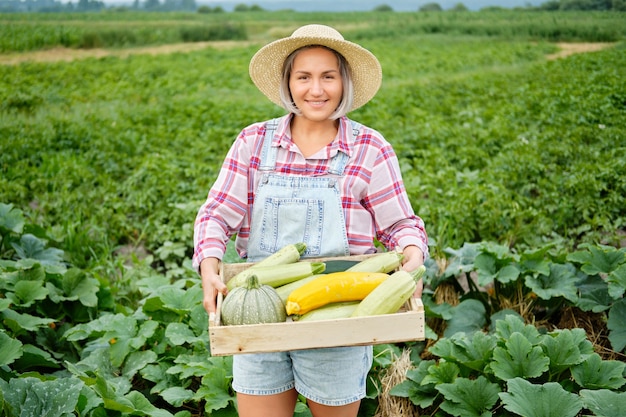 Симпатичная девочка в шляпе, собирающая свежие кабачки и кабачки в саду. Осенний урожай овощей.