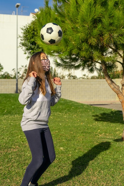 Милая девушка в маске играет в футбол на лужайке.