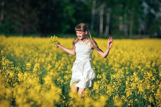 A cute girl in a white dress runs through a rapeseed field with a bouquet in her hands and laughs