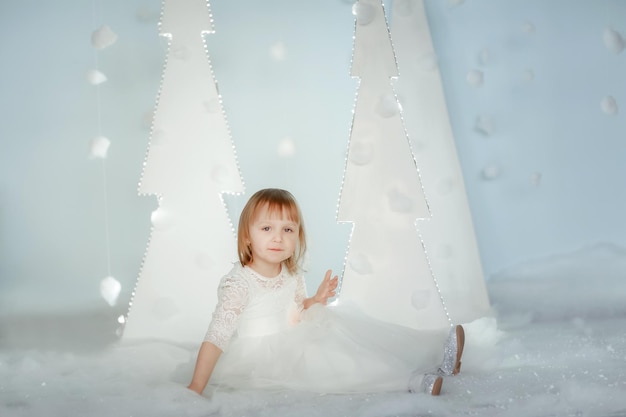 Cute girl in white Princess costume between white artificial glowing Christmas trees.