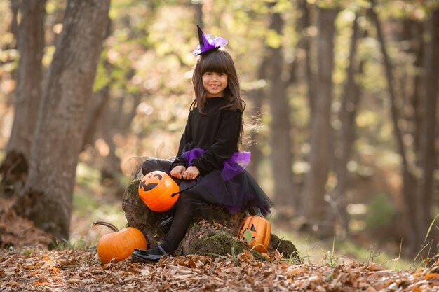 cute girl in witch costume is sitting on stump in autumn forest with pumpkin lantern Jack halloween