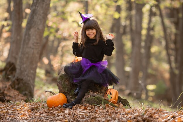 cute girl in witch costume is sitting on stump in autumn forest with pumpkin lantern Jack halloween