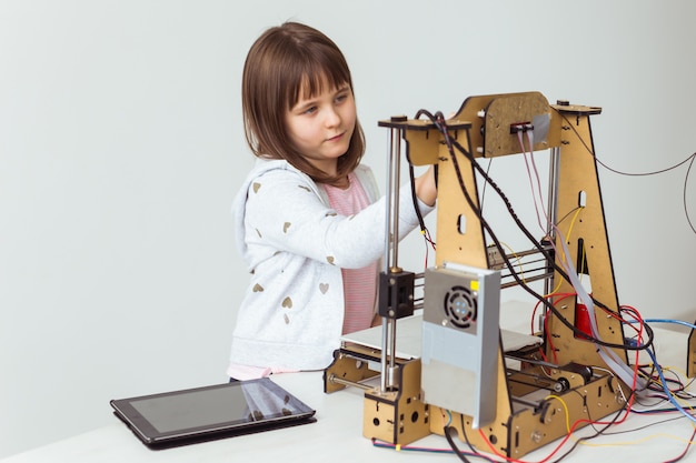 3Dプリントのシャッターシェードを備えたかわいい女の子は、3Dモデルをプリントする3Dプリンターを見ています。