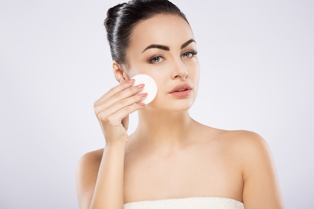 Cute girl with black hair fixed behind, big eyes, thick eyebrows and naked shoulders standing at gray studio background and holding cosmetic cleaning sponge, portrait.