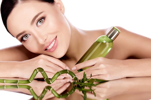 Cute girl with brown hair fixed behind, clean fresh skin, big eyes and naked shoulders posing with beauty product and leaves at white  wall.