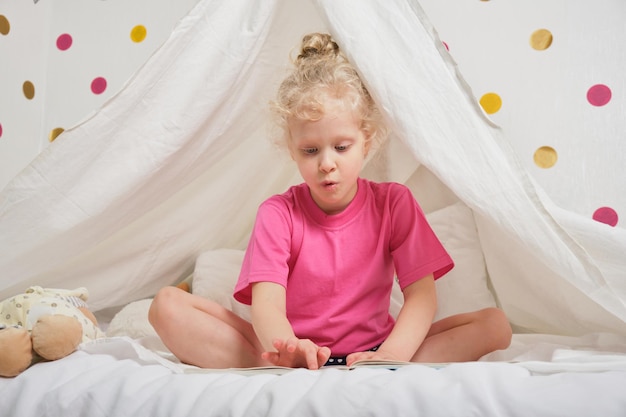 A cute girl with curly hair reads a book in a hut from a sheet on the bed, little girl having fun and playing in her tent, Hut in the kids room