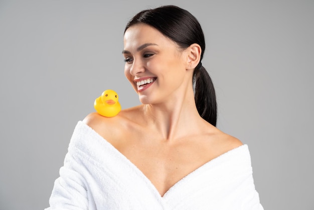 Cute girl with dark hair and light nude make-up wearing white bath robe, looking at the rubber duck and smiling over the white studio background. Beauty photo concept