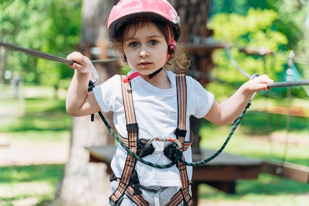Cute girl with fear in her eyes looks straight into the camera. A small child in a safety helmet and insurance stands on a rope