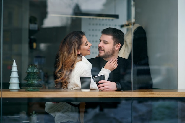 Cute girl with her boyfriend sitting inside a cafe and drinking hot fragrant coffee while outside is cold weather. Love and romance .