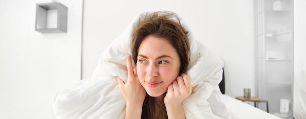 Foto una ragazza carina con i capelli disordinati sdraiata a letto coperta di lenzuola bianche, sorridente e ridendo coquette