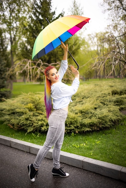 A cute girl with multi-colored braids and bright makeup in a bluish shirt posing with a rainbow umbrella against the background of a blooming spring park