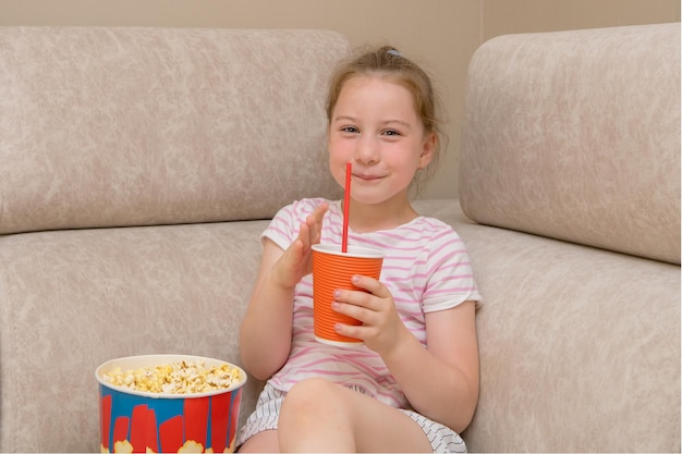 Cute girl with a pigtail joyfully drinks cola and eats popcorn while sitting on the couch at home