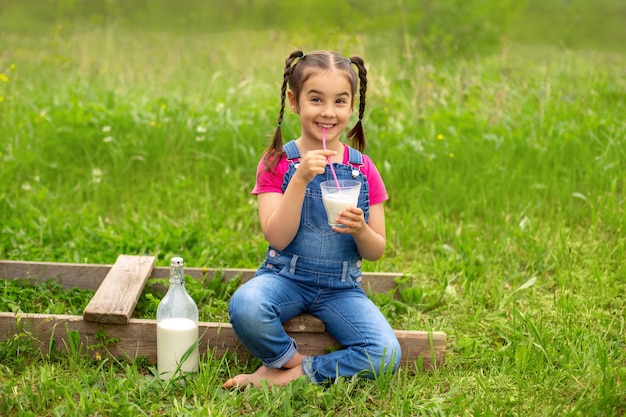 Cute girl with pigtails, holds a glass of milk with a pink straw, sits on a green lawn