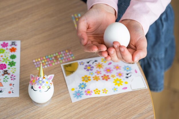 Photo a cute girl with pink bunny ears makes an easter craft decorates an egg in the form of a unicorn with rhinestones horn flowers in the interior of a house with plants