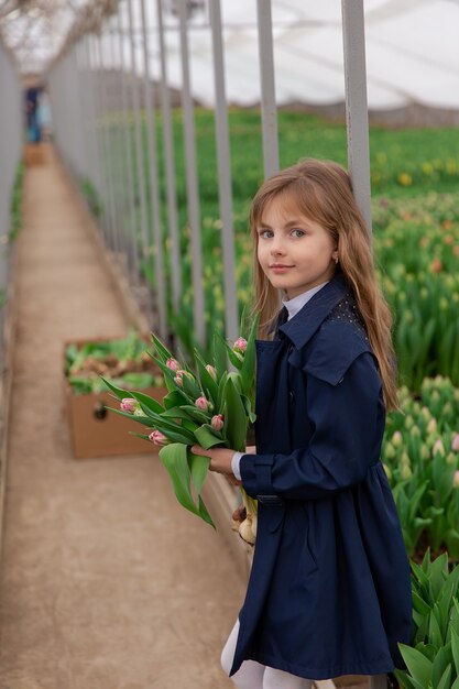 Ragazza carina con tulipani con i capelli biondi in primavera in una serra con tulipani in crescita