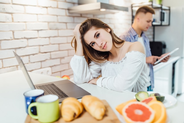 Cute girlfriend doing work on the laptop while man searching recipe for dinner