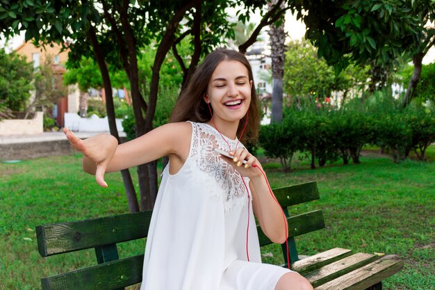 Cute gorgeous woman is listening to music in headphones and dancing on the bench in the park in a warm day