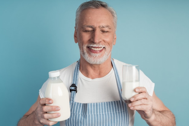 Cute grandfather with gray hair and beard holds a bottle of milk and a glass, wears an apron