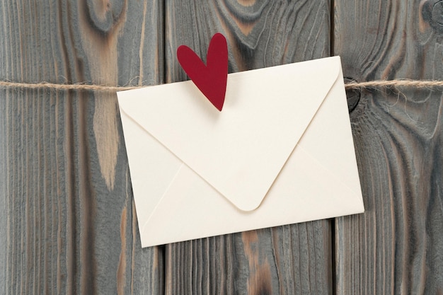 Симпатичный конверт из белой бумаги ручной работы с красным сердцем в виде штампа, висящего на веревке на темном деревянном фоне. Секретный подарок