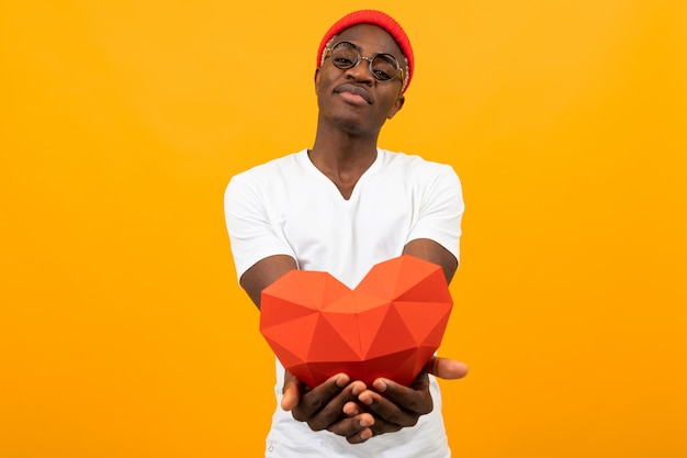 Cute handsome American in a white T-shirt holds out a red 3D heart made of paper for Valentine's Day on yellow with copy space