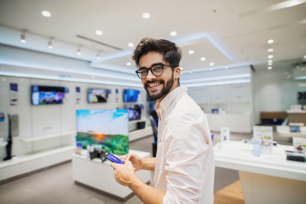 Cute handsome young man standing in bright electronic store and looking at the camera. Testing some new phones.