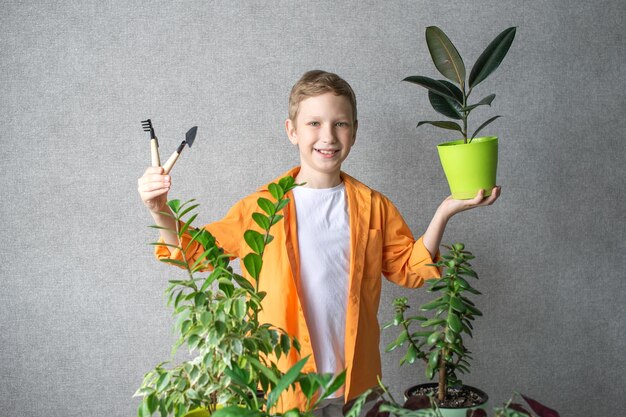 シャツを着たかわいい幸せな農学者の少年が、屋内の植物と土壌管理用の道具を持って立っています