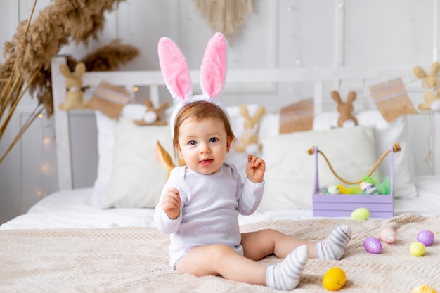 cute happy baby girl in bunny ears on the bed at home with a rabbit and painted eggs easter concept funny Easter baby is waiting for the holiday and rejoicing