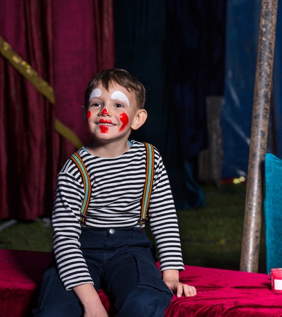Милый счастливый мальчик с клоунским макияжем сидит на сцене перед представлением в полосатой рубашке с длинным рукавом и подтяжками