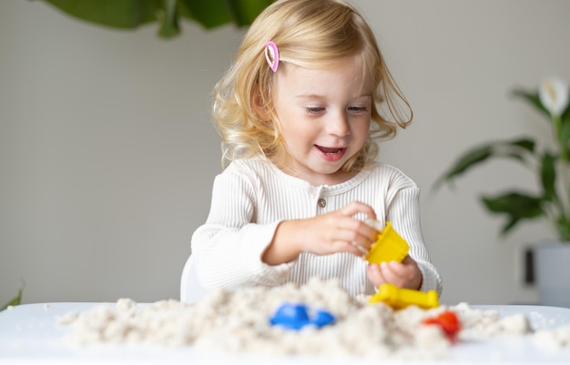 Cute happy caucasianblondecurlyhaired toddlerbaby girl playing with kinetic sand indoorsPreschool kid early developmentmotorsensoric skills concept