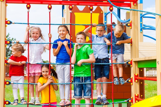 Cute and happy friends of different age climbing on rope net as part of outdoor playground. Kids having fun posing on climbing net